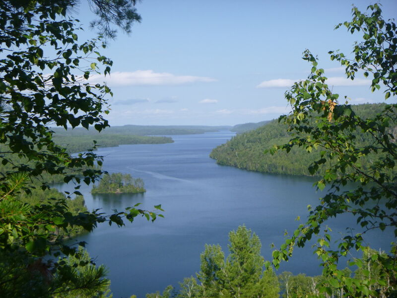 The image captures a serene view of a wide, calm river or lake, framed by lush green trees on either side. A small, tree-covered island sits in the middle of the water. The sky is blue with a few scattered clouds, contributing to the peaceful and natural scenery. The composition emphasizes the tranquility and beauty of the landscape.